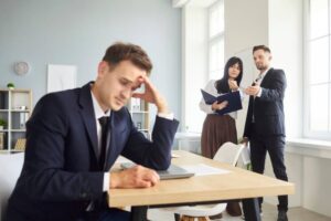 Man at office desk rests head in hand as peers talk by window, with workplace stress, gossip, conflict, bullying